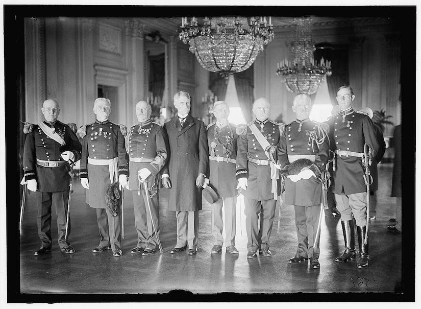 Eight men, seven of whom wear Army dress uniforms, stand in a line in a reception room at the White House.