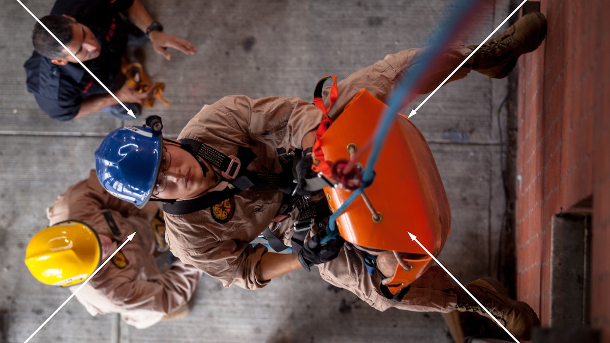 Military rescuers send a rescue toboggan up a wall. The main subject is in the center and is looking up.