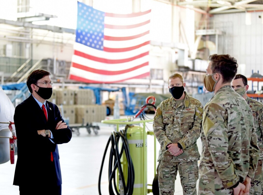 A man dressed in civilian clothing stands facing three men wearing military uniforms in a hangar with a U.S. flag hanging from the ceiling. All are wearing face masks.