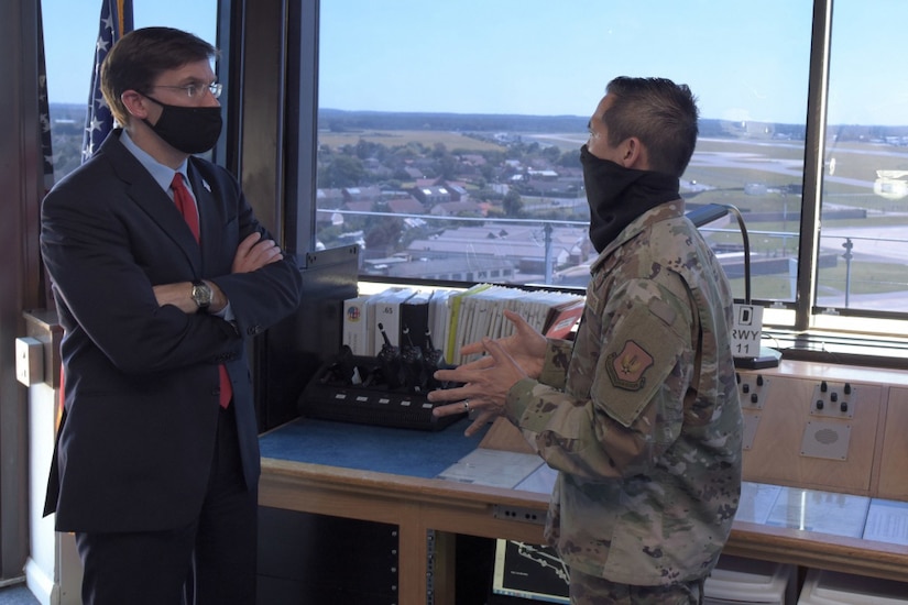 Two men wearing face masks, one dressed in a business suit and one dressed in a military uniform, talk in a small office next to a window overlooking the countryside.