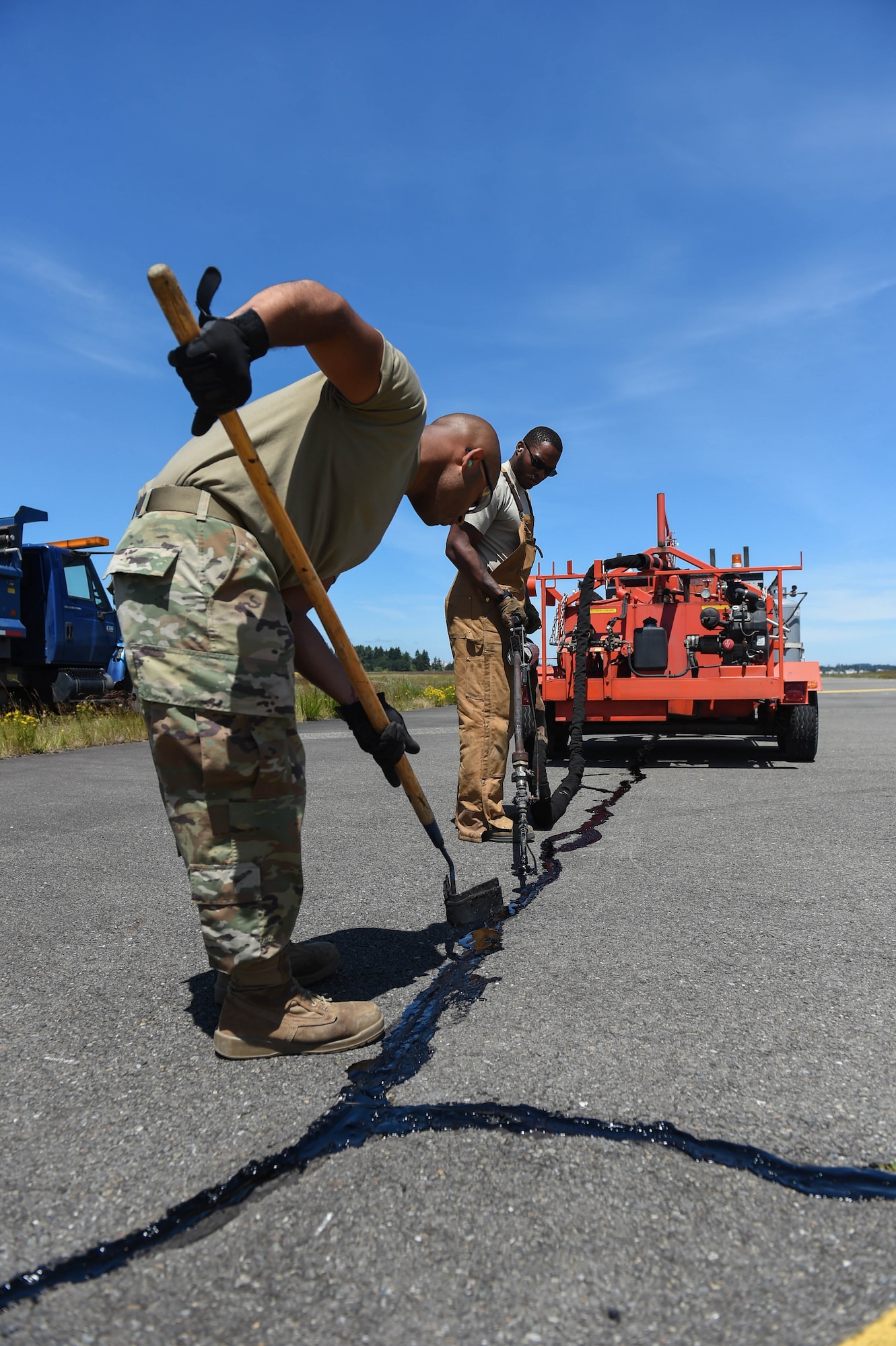 Airman 1st Class Felipe Reyes-Cedeno, left, and Senior Airman Irvin Matthews, right, 627th Civil Engineer Squadron pavements and construction equipment technicians work together to reseal a crack in the McChord Field flight line on Joint Base Lewis-McChord, Wash., June 25, 2020. In a three-person resealing team, one person drives the truck moving the tar kettle around, while another operates the tar dispensing handle, and the last person follows them with a squeegee making sure the tar is level with the asphalt. (U.S. Air Force photo by Airman 1st Class Mikayla Heineck)