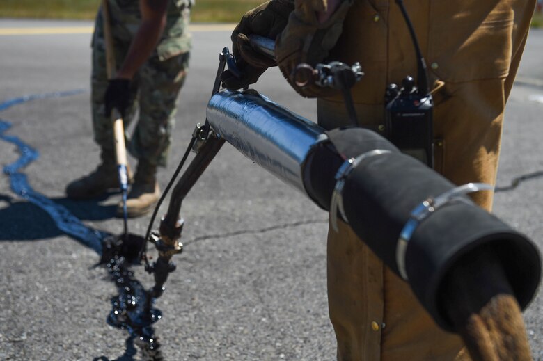 Airman 1st Class Felipe Reyes-Cedeno, left, 627th Civil Engineer Squadron pavements and construction equipment technician, uses a squeegee to level melted tar with the asphalt as Senior Airman Irvin Matthews, right, 627th CES pavements and construction equipment technician, dispenses the tar into the cracks on the McChord Field flight line on Joint Base Lewis-McChord, Wash., June 25, 2020. Filling pavement cracks helps prolong its life by preventing rain and moisture from flowing through the asphalt and eroding it. (U.S. Air Force photo by Airman 1st Class Mikayla Heineck)