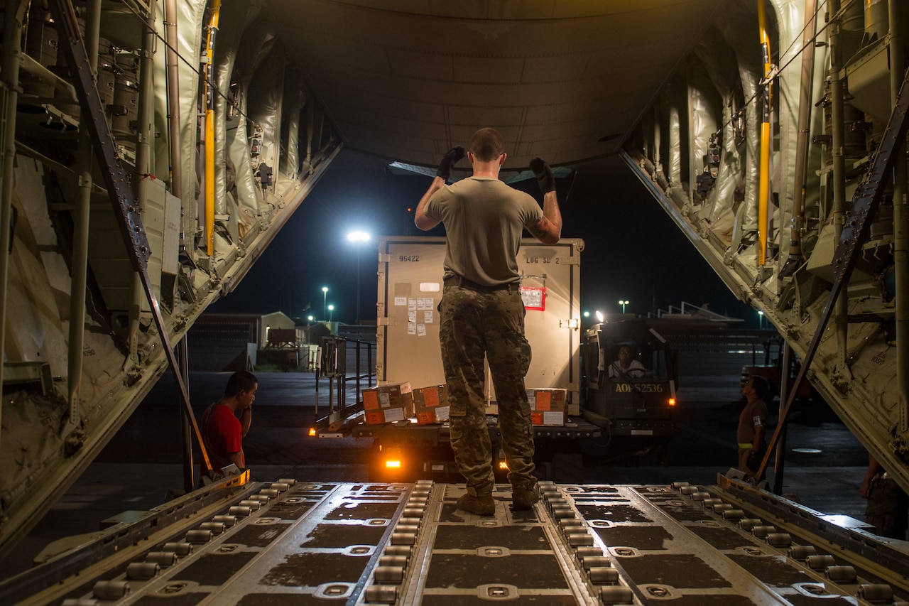 A person in uniform guides a truck as it backs up to the cargo loading area of an airplane.