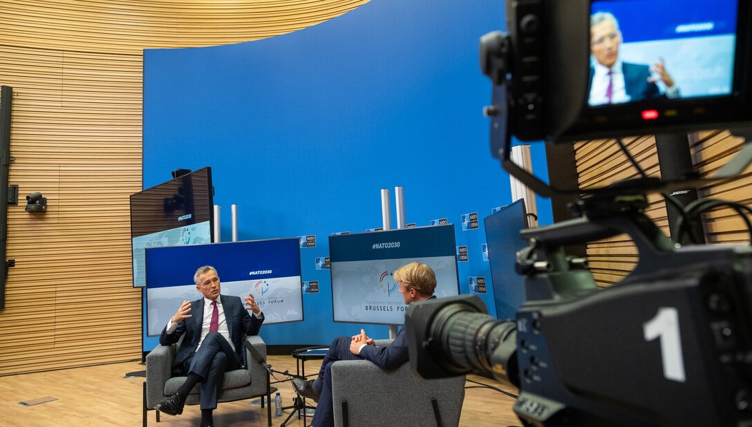 Two men speak together on the set of a television studio. There is a television camera in the foreground.