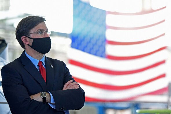 A man dressed in civilian clothing stands facing three men wearing military uniforms in a hangar with a U.S. flag hanging from the ceiling. All are wearing face masks.