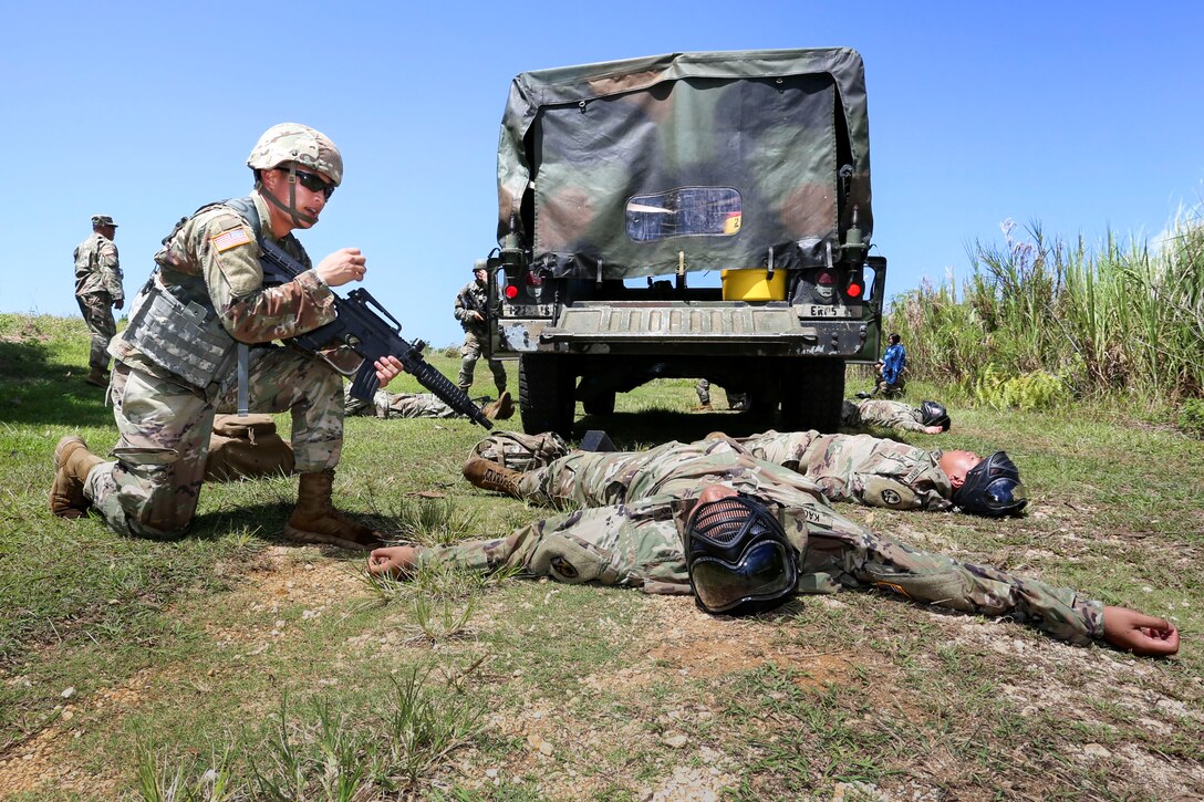 Two officer candidates lie on their backs behind a truck in a field, while one, holding a rifle, kneels beside them.