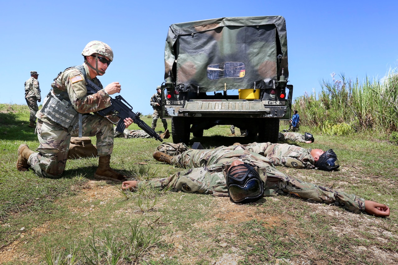 Two officer candidates lie on their backs behind a truck in a field, while one, holding a rifle, kneels beside them.