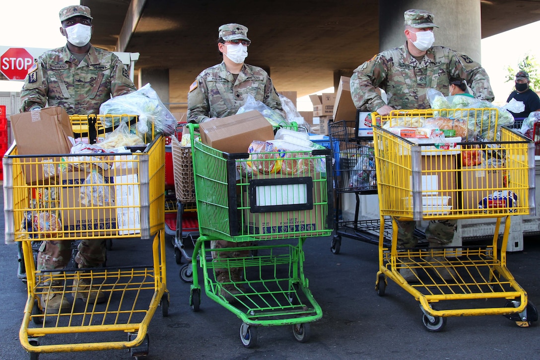 Three service members wearing face masks and gloves prepare to deliver grocery carts filled with food to waiting vehicles.