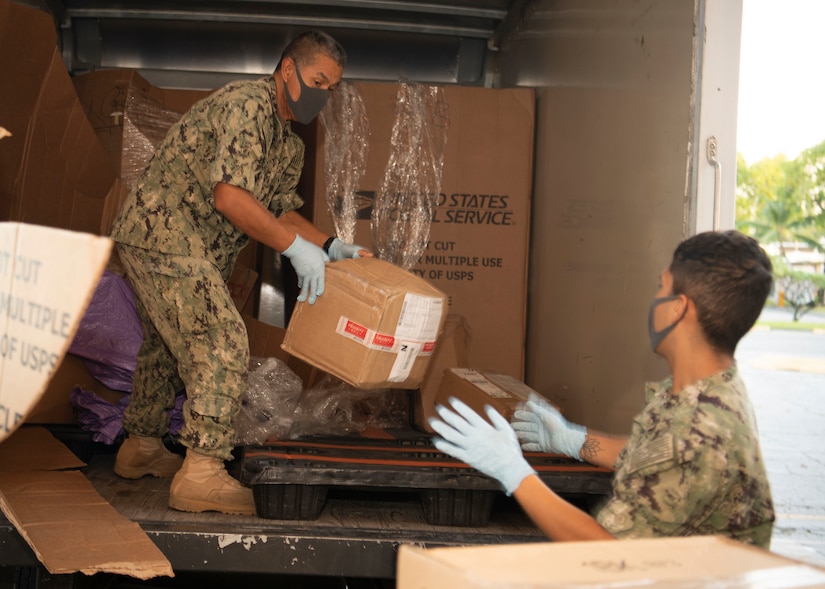 A sailor wearing a face mask unloads a box from a truck and gives it to another sailor, who is also wearing a face mask.