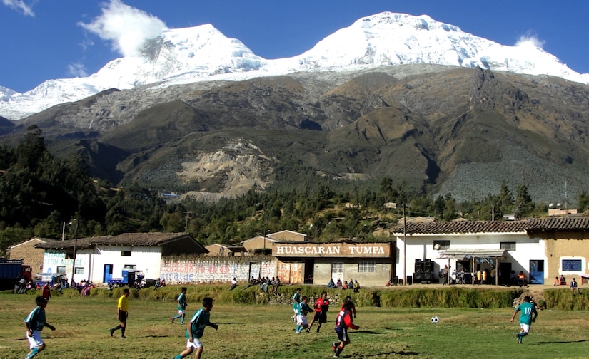 Soccer players run on a field with small buildings along the edge. Snow-capped mountains loom in the background.