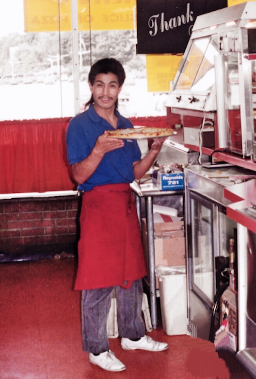 A man wearing an apron holds a pizza in a pizza shop.