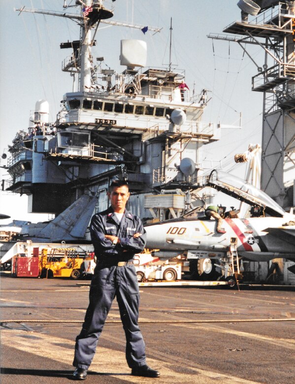 A sailor stands with arms crossed on the deck of an aircraft carrier.
