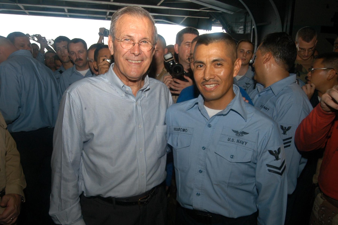 A sailor in uniform and a man dressed in civilian clothing pose for a photo while other sailors watch from behind.