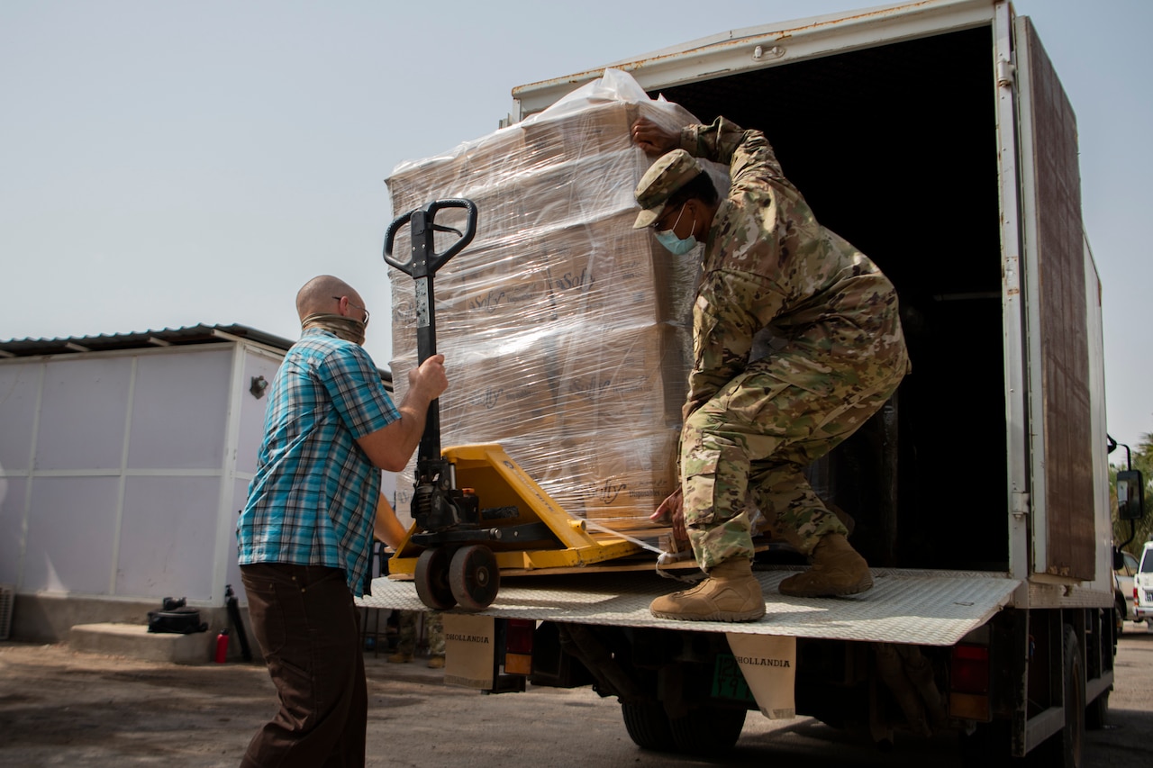Two soldiers — one in civilian clothes, one in uniform, wear face masks while unloading a pallet of supplies from a truck.