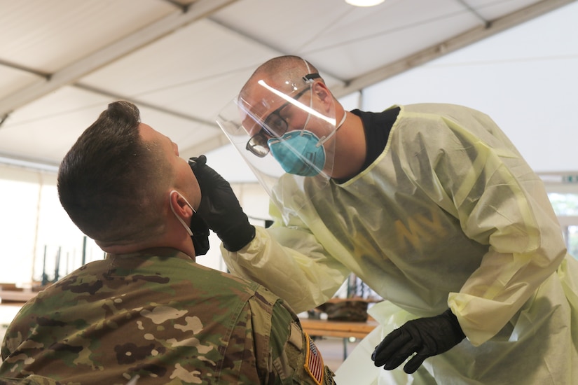 A medic wearing a face mask and plastic face shield administers a COVID-19 test to a soldier.