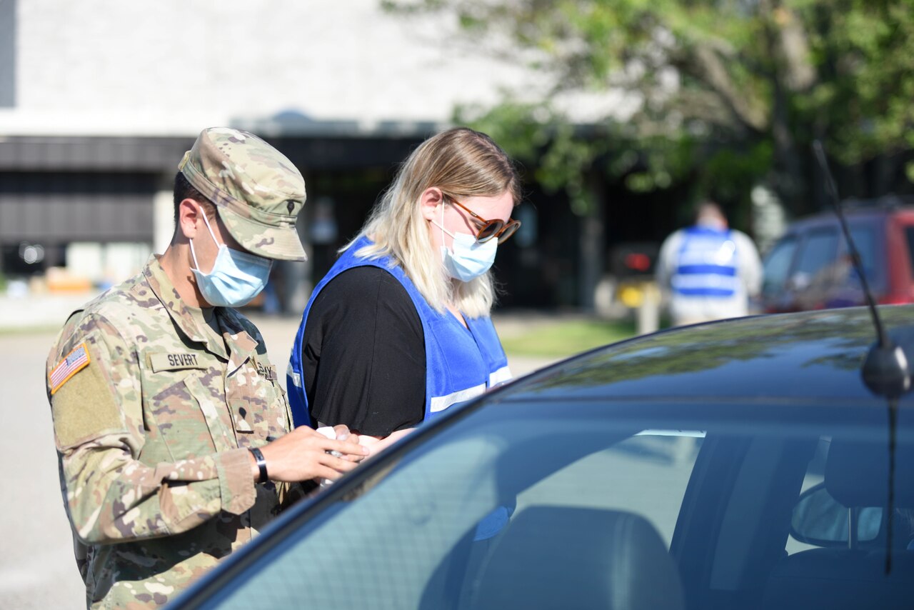 A soldier and a woman in civilian clothes conduct drive-thru COVID-19 testing.