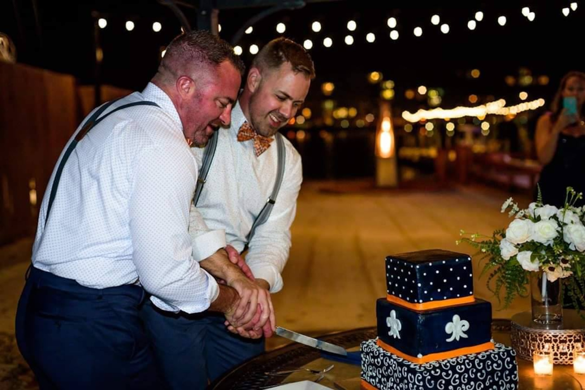 Two men stand next to each other at their wedding. Both are wearing white button-up shirts with suspenders and bowties. It's night in a festive atmosphere and they're slicing into their wedding cake.