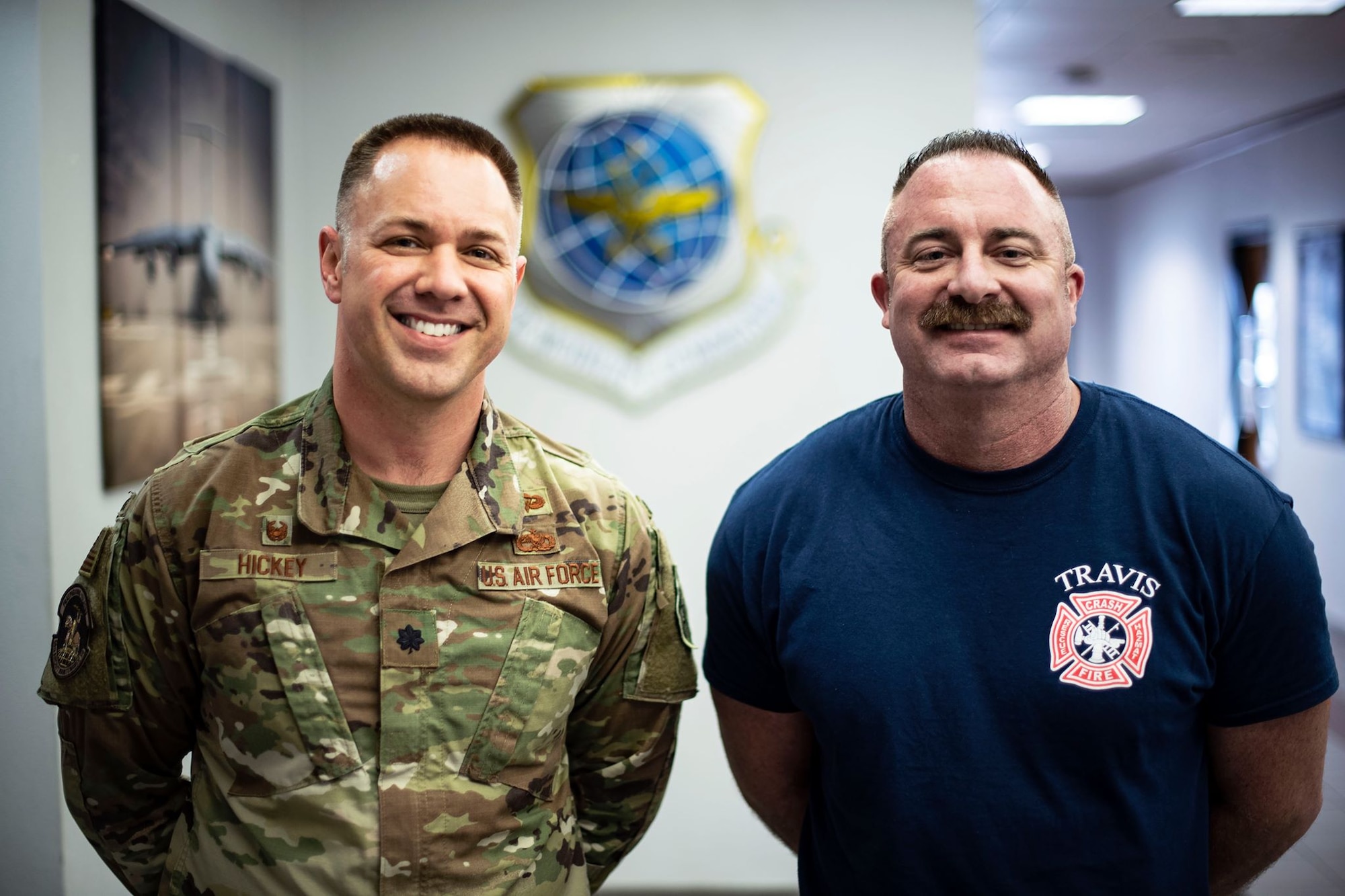 Two men stand next to each other inside of a brightly-lit building. One wears a military uniform and one wears a tee shirt with "Travis fire department" on it. Both are smiling.