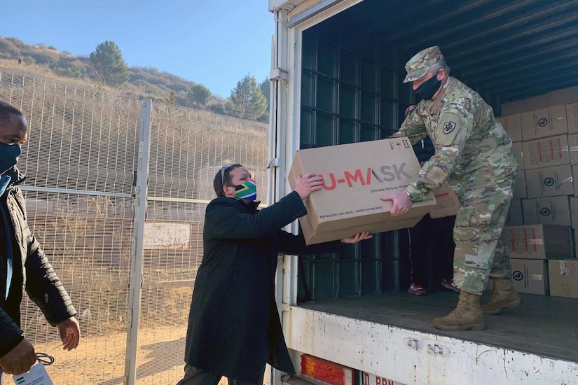 A soldier standing in a truck wearing a mask passes a box to someone standing on the ground and wearing a mask.