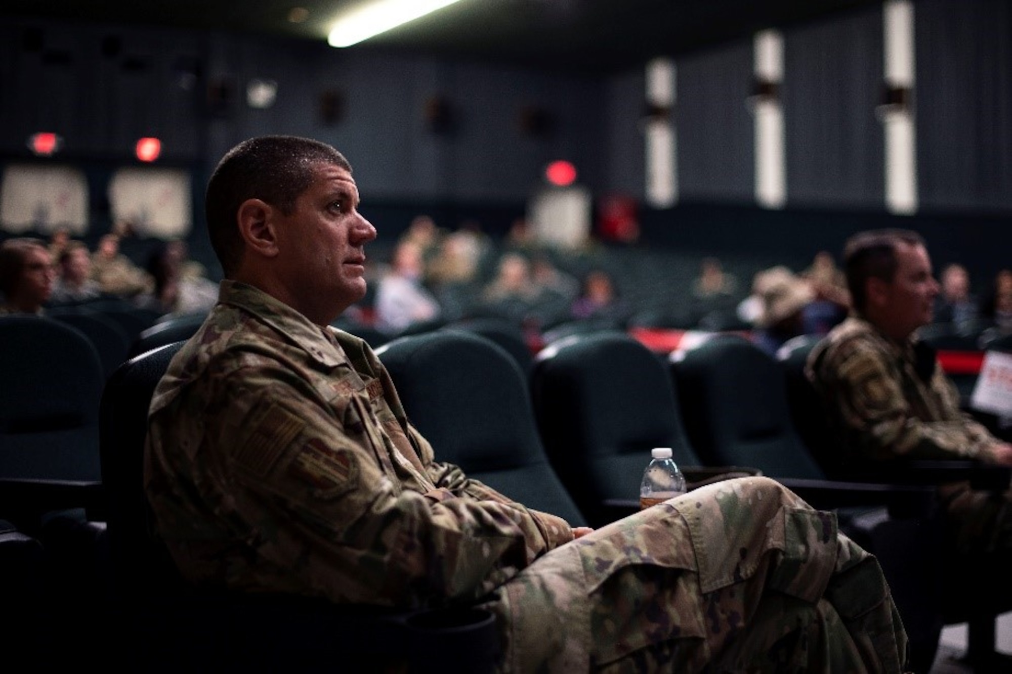 A man in a military uniform sits in a theater with others in attendance while he looks on at someone off screen.