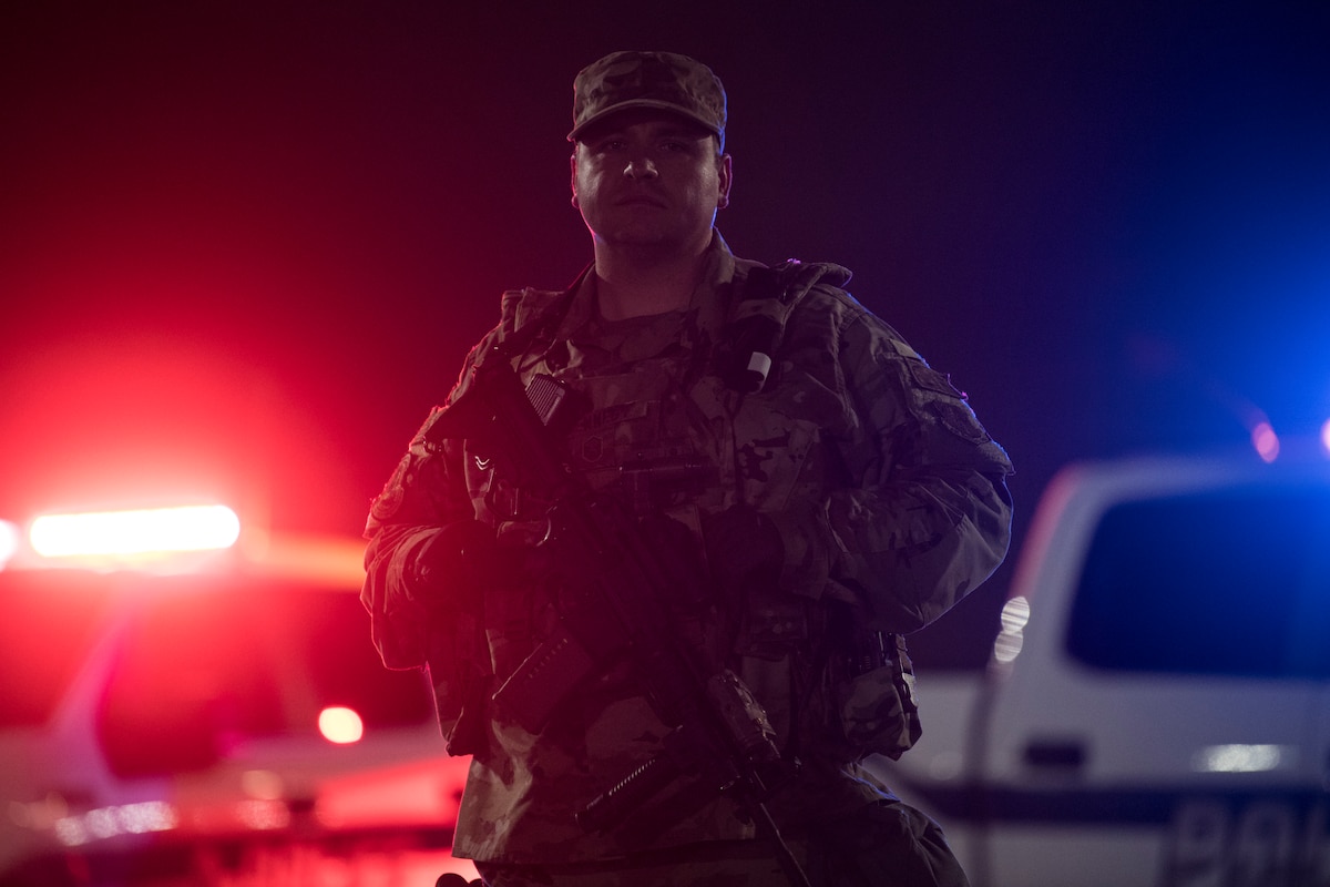 A photo of a U.S. Air National Guard Security Forces defender standing in front of two police vehicles that are flashing red and blue lights.