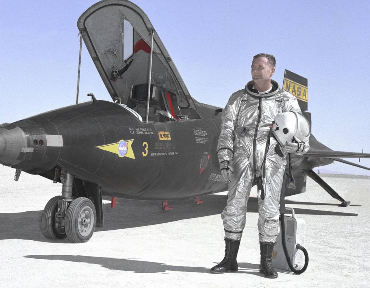 A man in a silver suit stands in front of a military-style research aircraft.
