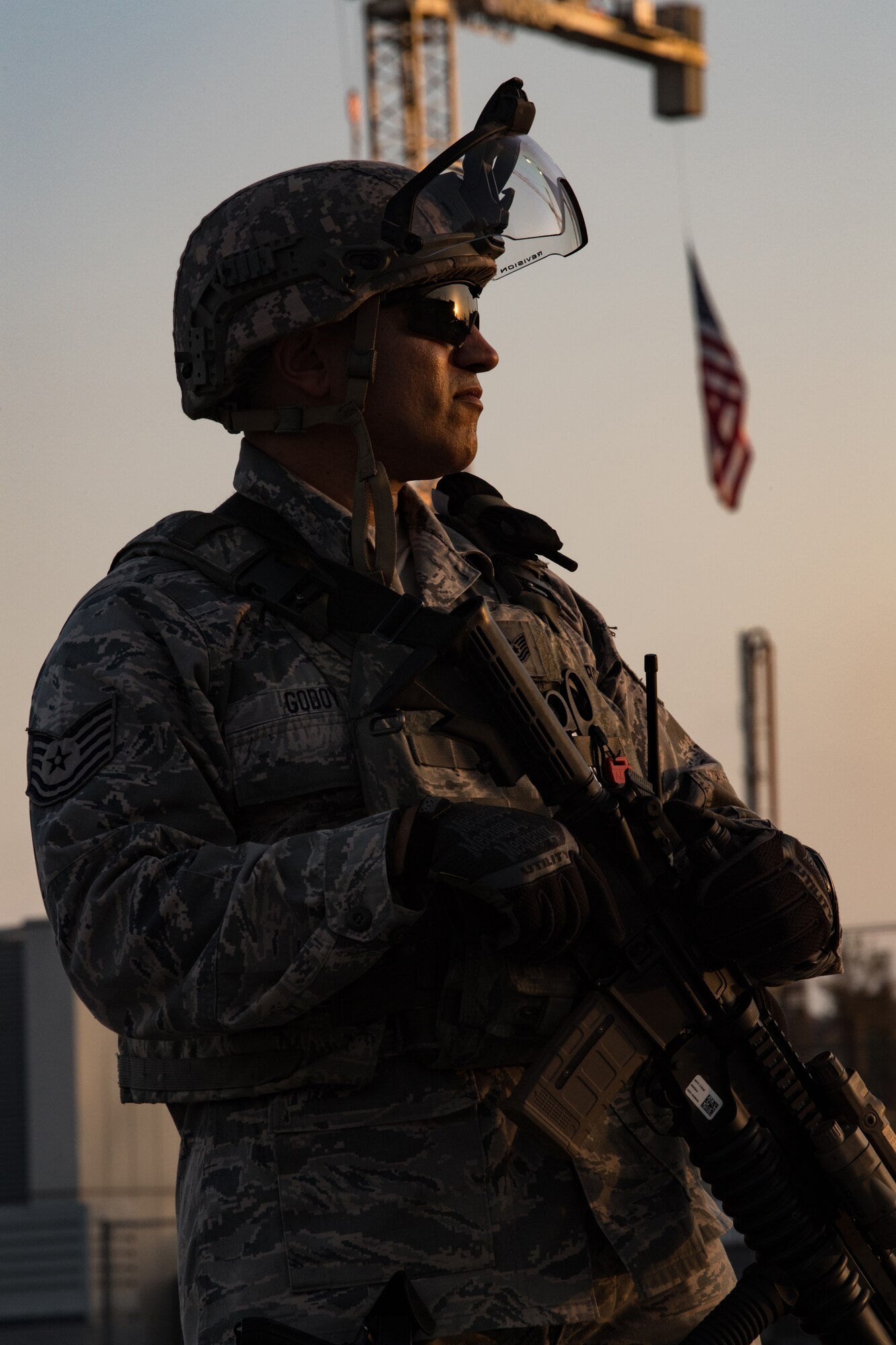 A California Air National Guard Security Forces defender stares into the sunset as the outline shape of his body silhouettes from the setting sun. A U.S. Flag hangs from a construction crane behind him.