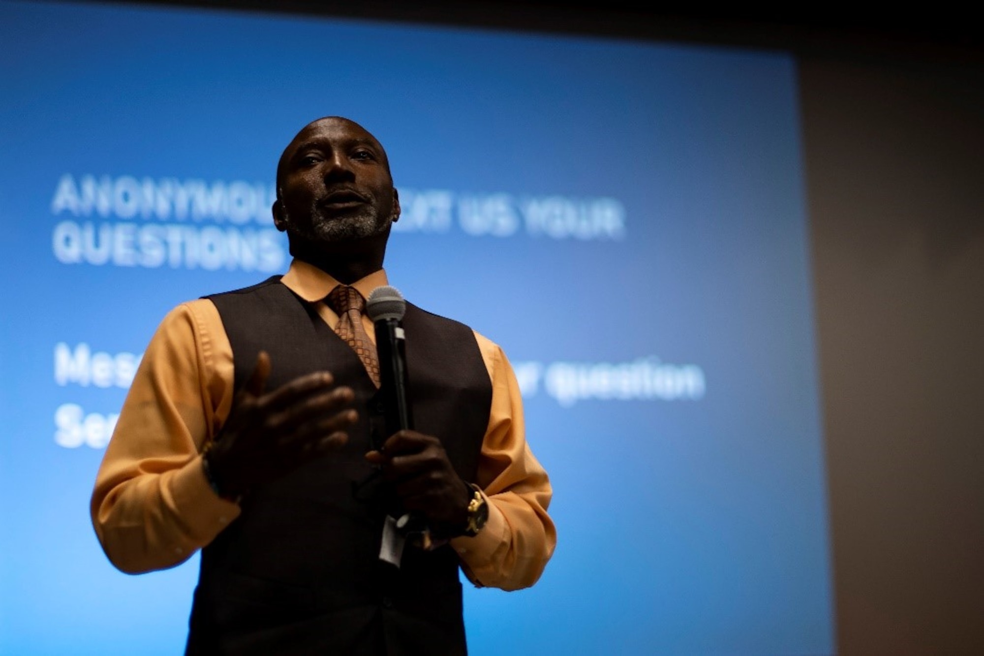 A well-dressed man holds a microphone while he addresses an audience off-screen.