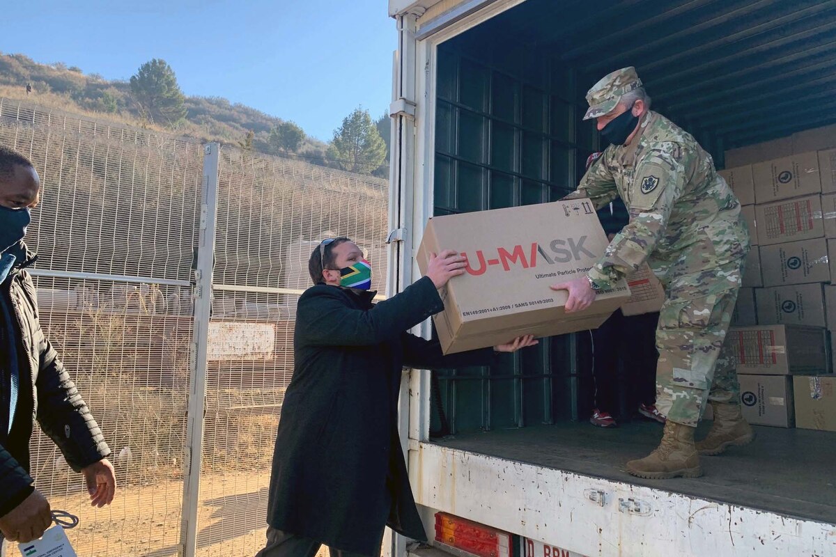 A soldier standing in a truck wearing a mask passes a box to someone standing on the ground and wearing a mask.