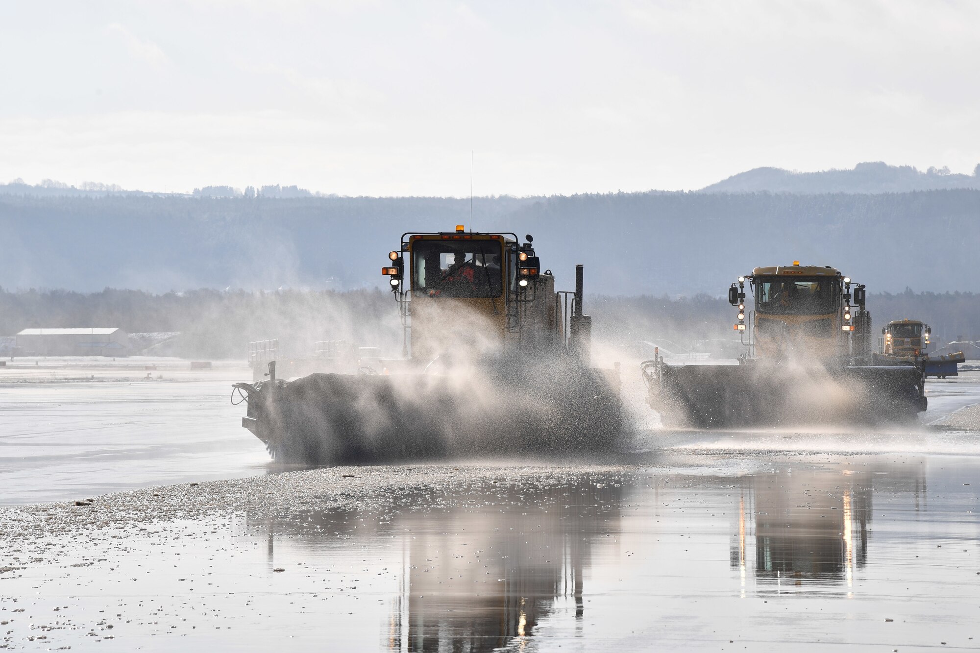 Members of the 786th Civil Engineer Squadron snow operations team drive Oshkosh Snow Broom snow removal vehicles across an airfield ramp to sweep away remaining snow and ice after winter weather on Ramstein Air Base, Germany, Feb. 28, 2020.