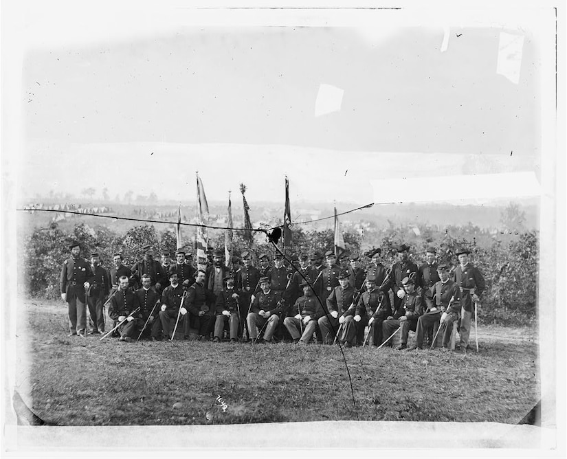 A unit of men in Civil War garb pose for a photo in front of a crop field.