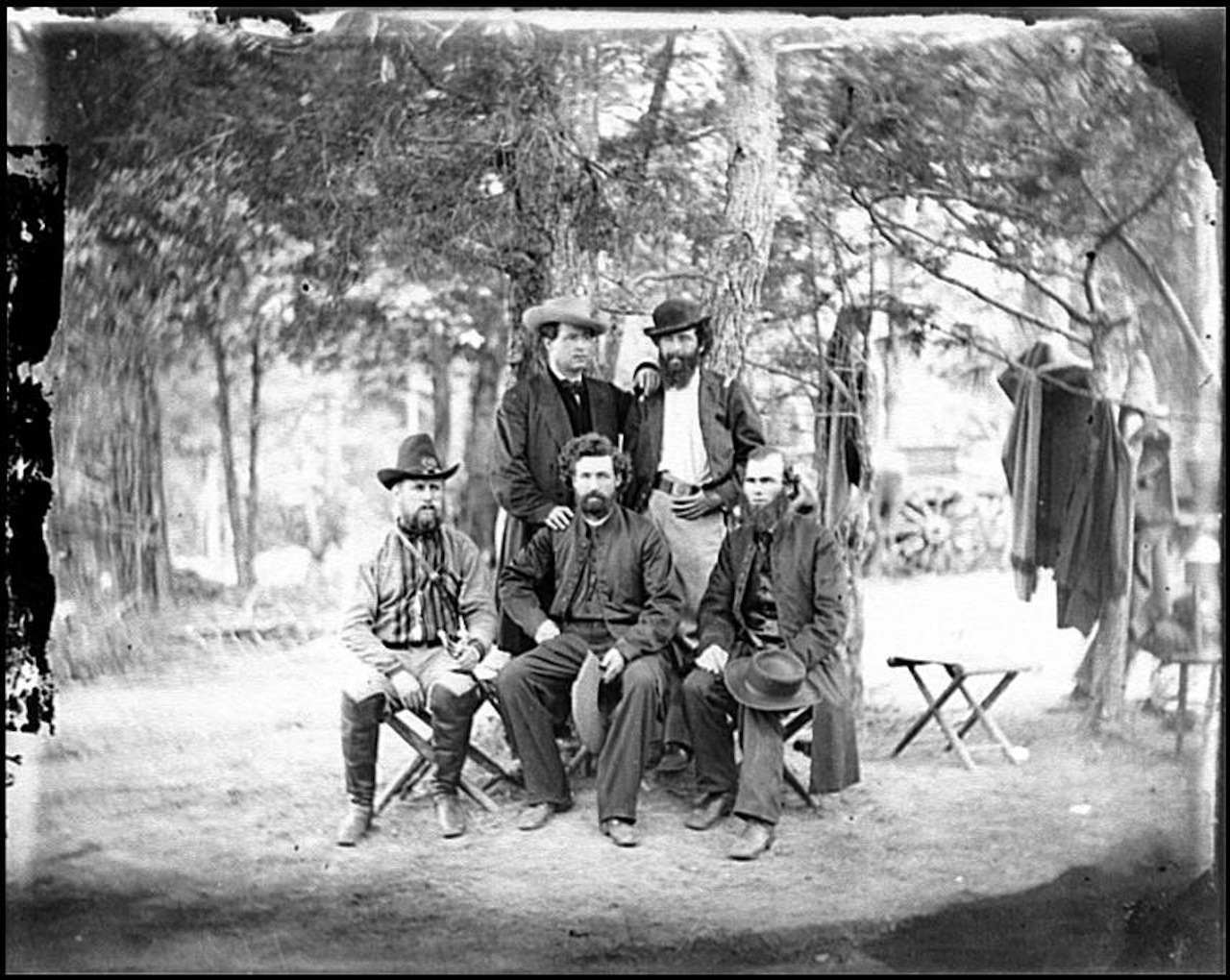 Five Civil War-era men, two standing and three sitting, pose for a black-and-white photo in a wooded area. Clothes can be seen hanging from tree limbs nearby.