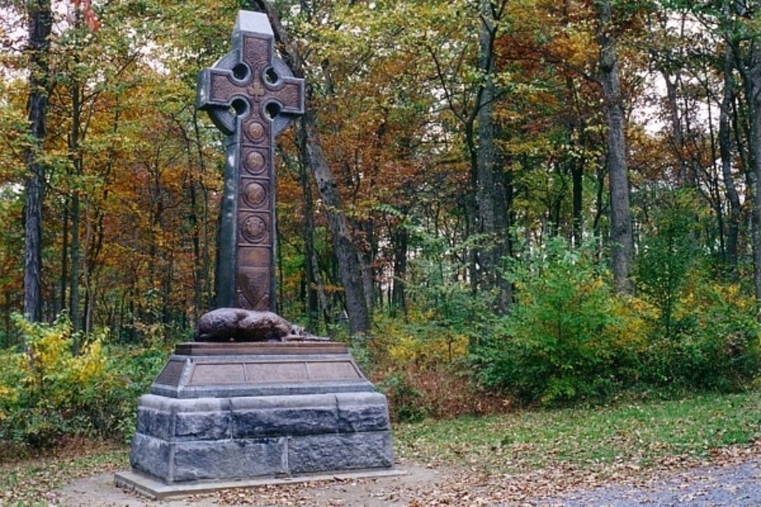 A large cross-shaped monument with a bronze wolfhound at its base sits along a path with woods behind it.