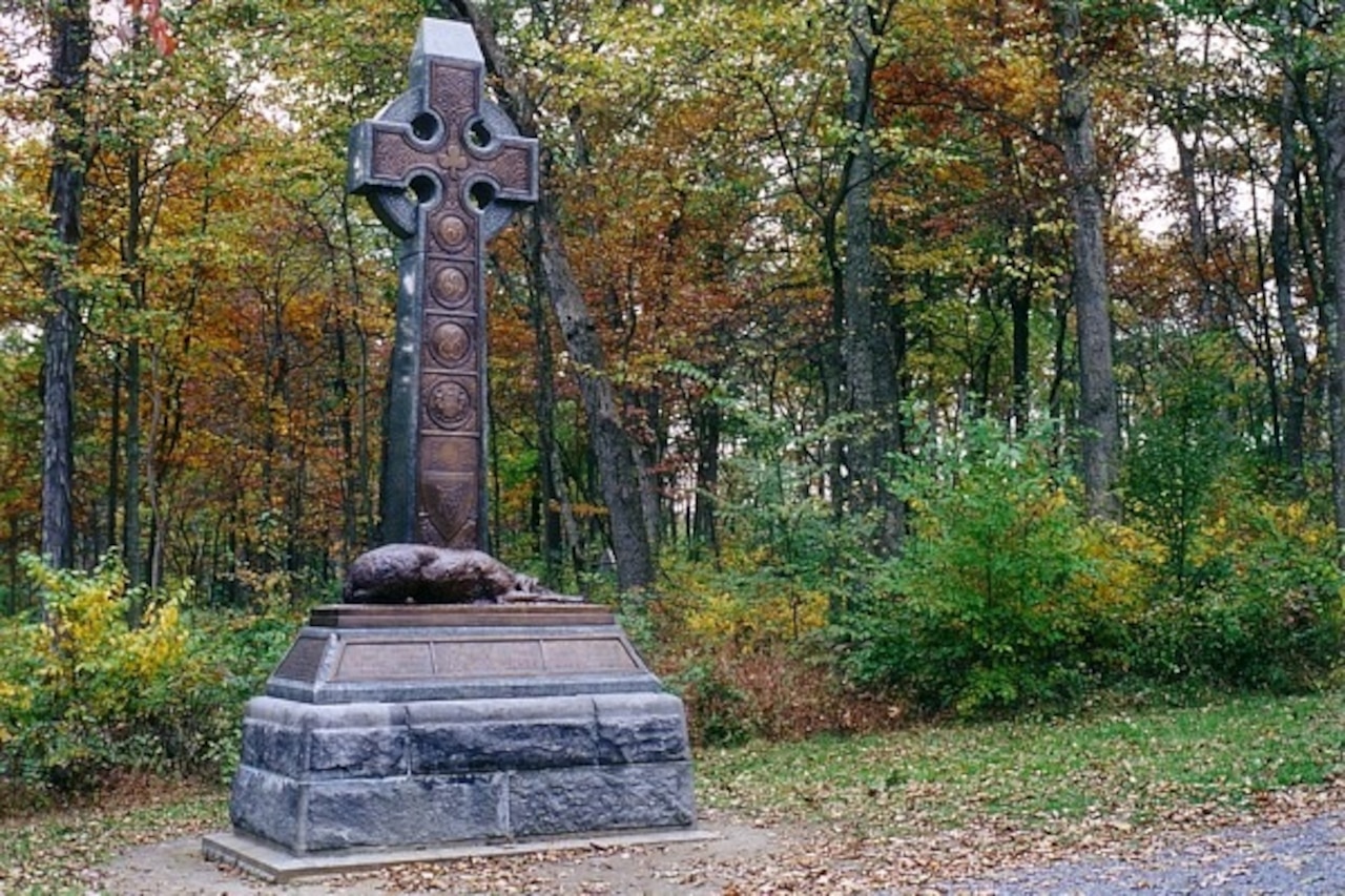 A large cross-shaped monument with a bronze wolfhound at its base sits along a path with woods behind it.