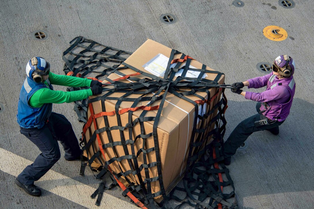 Two sailors untie a net surrounding a large box.