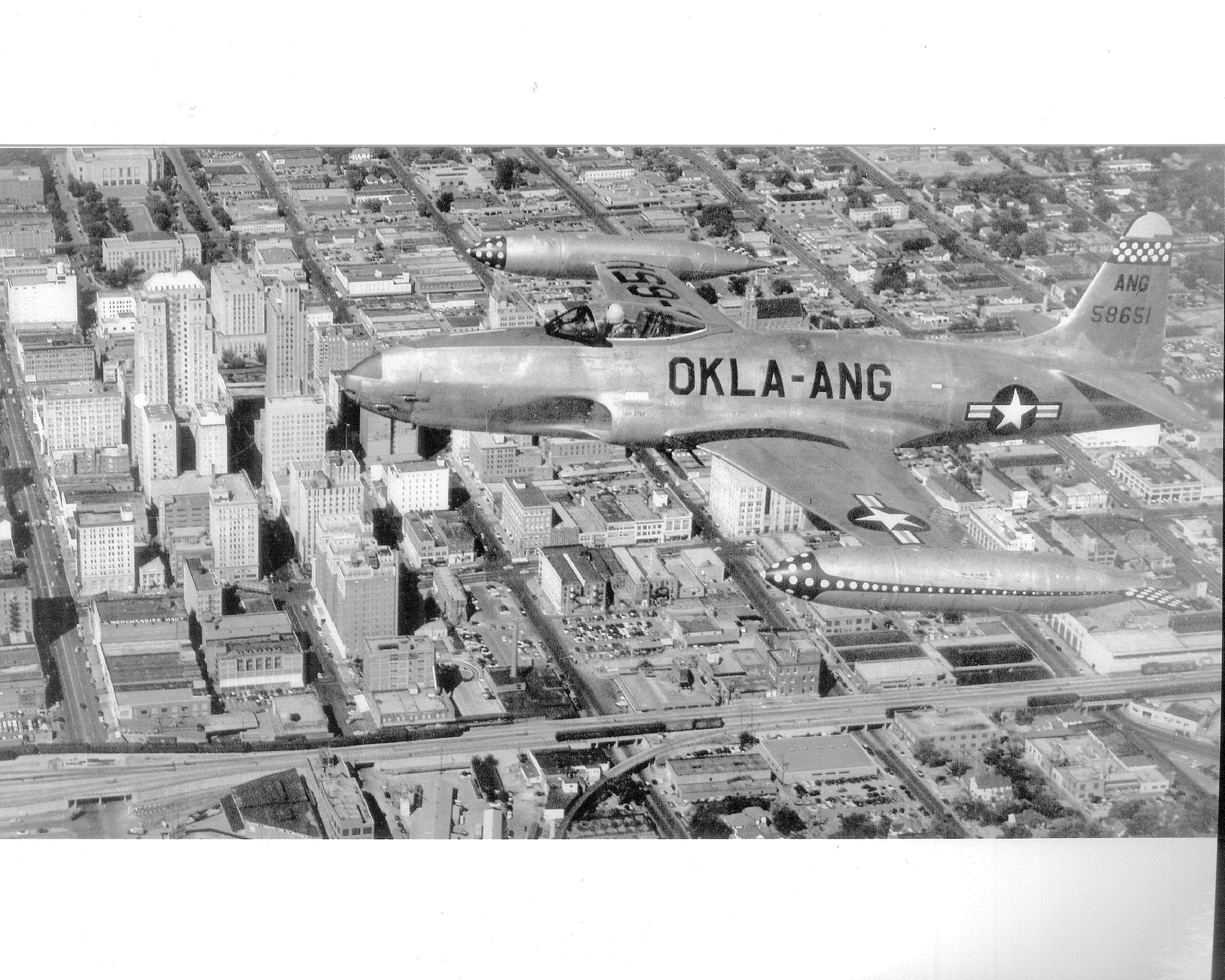 Black and white aerial photo of F-80 Shooting Star flying over a city.