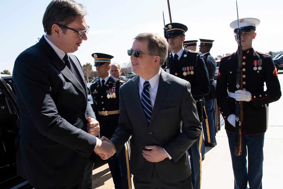 Two men shake hands while men in uniform stand behind them.