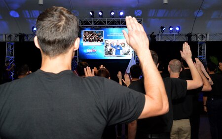 Males in black t-shirts with right hand raised face a tv screen with man in blue jumpsuit raising his right hand.