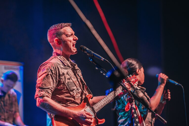 A soldier sings and plays guitar as another holds a microphone in the background.