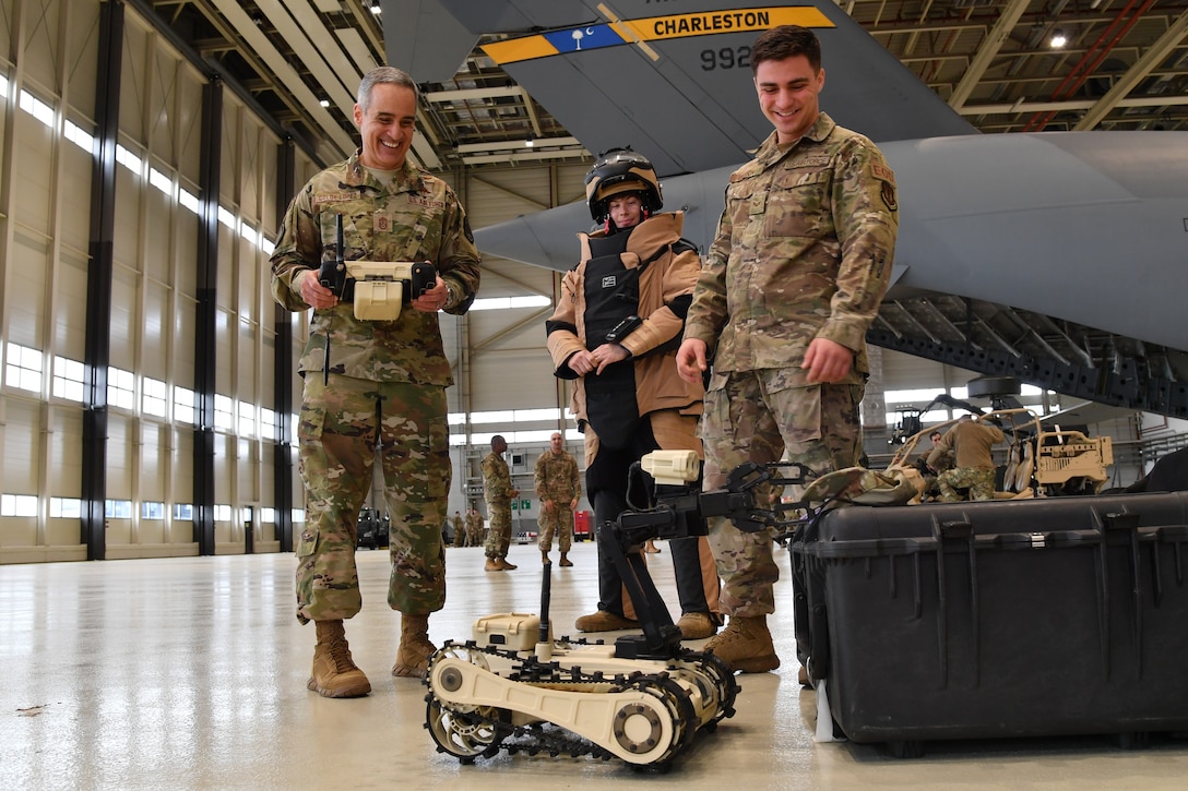 Man in uniform uses remote control to maneuver robot as two others watch.
