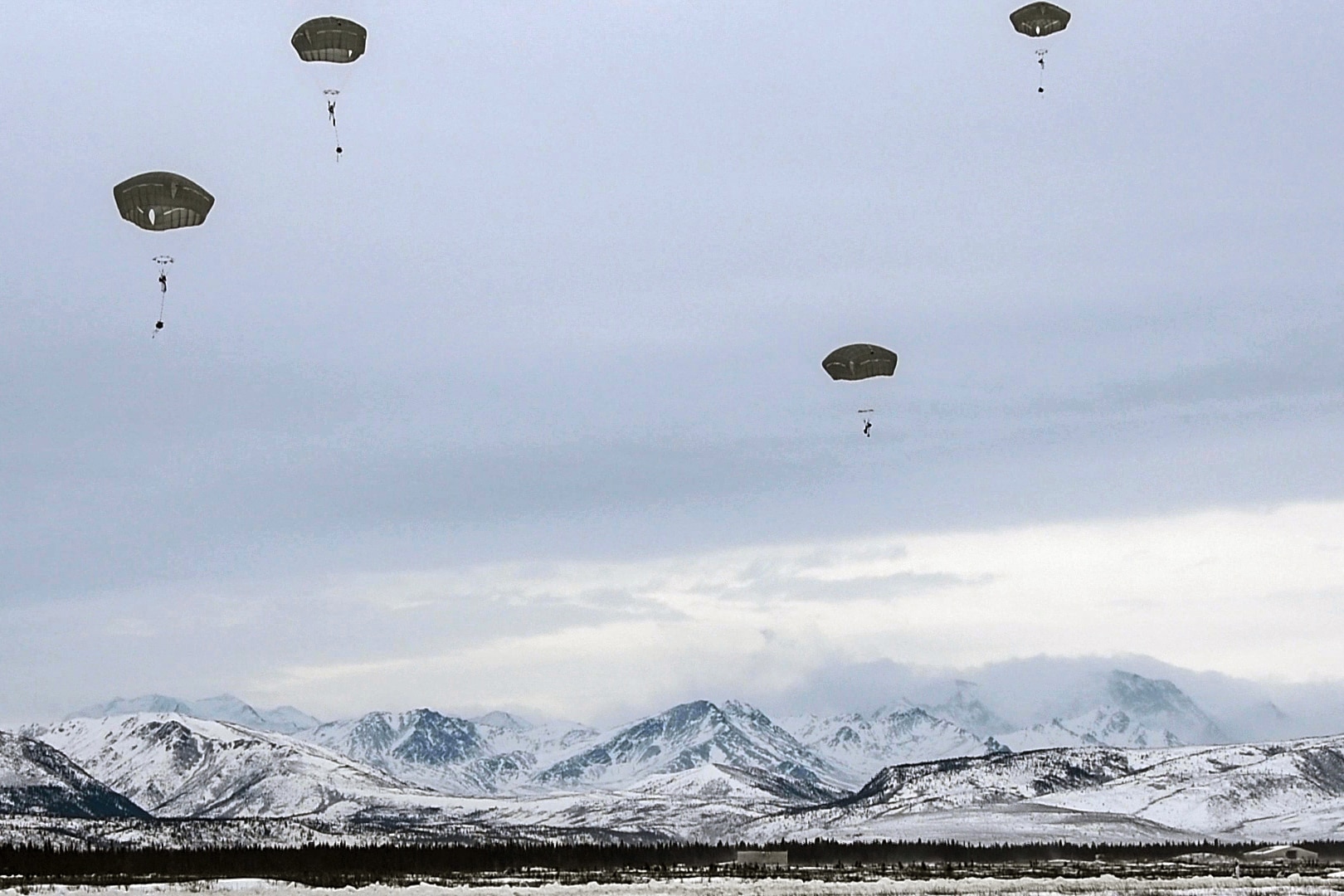 Parachutists drop down onto a snowy landscape with mountains in the background.