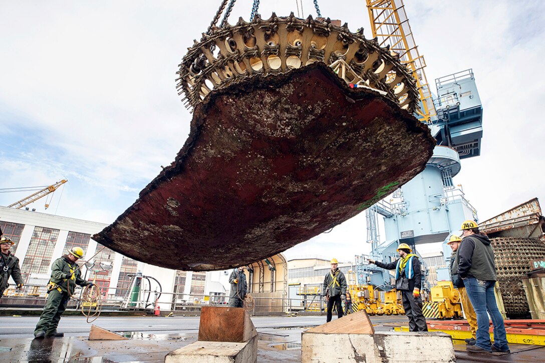 Riggers watch as a crane lifts a massive piece of submarine hull onto a barge for disposal.