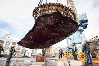 Riggers watch as a crane lifts a massive piece of submarine hull onto a barge for disposal.