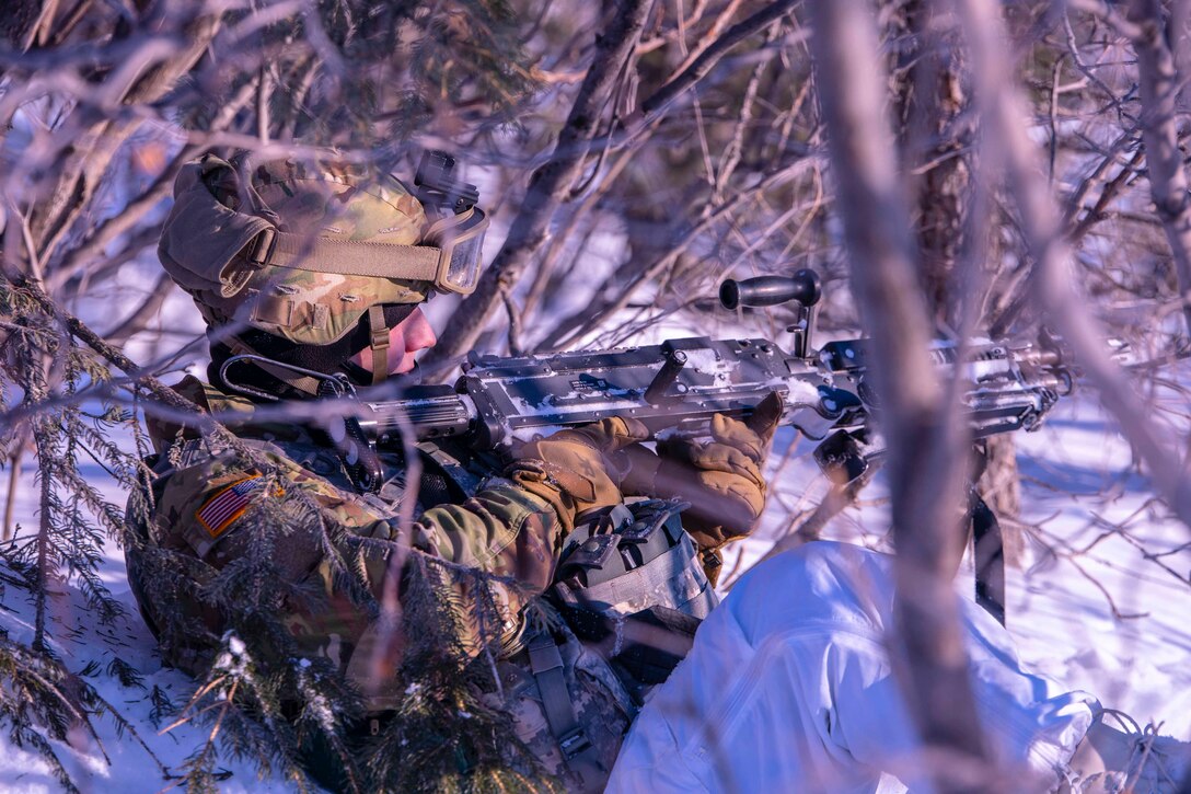 A soldier sits in snow surrounded by thin trees while pointing a weapon.