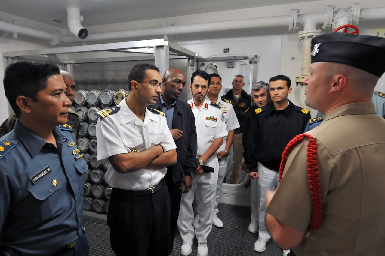 A group of military personnel, dressed in a variety of uniforms, stand next to each other while listening to another service member speak.