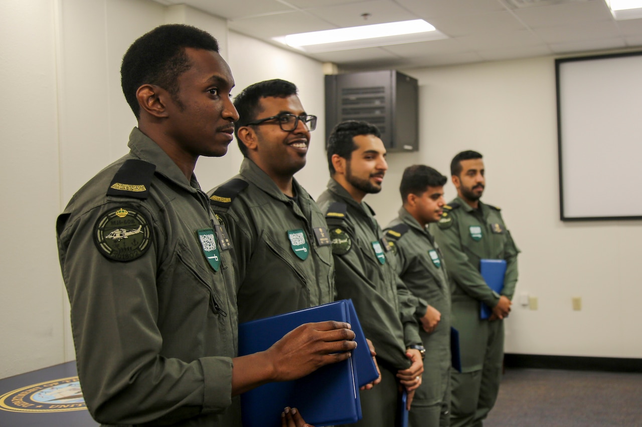 Five military service members stand in a line; each holds a blue folder.
