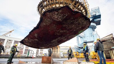 Riggers watch as a crane lifts a massive piece of submarine hull onto a barge for disposal.