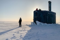 A service member stands on ice as other service members watch from a submarine.