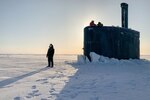 A service member stands on ice as other service members watch from a submarine.