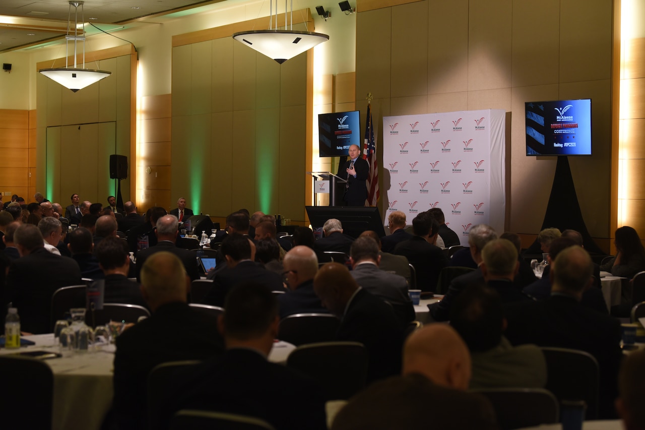 People seated at tables in a big room listen to a man speaking at the front of the room.